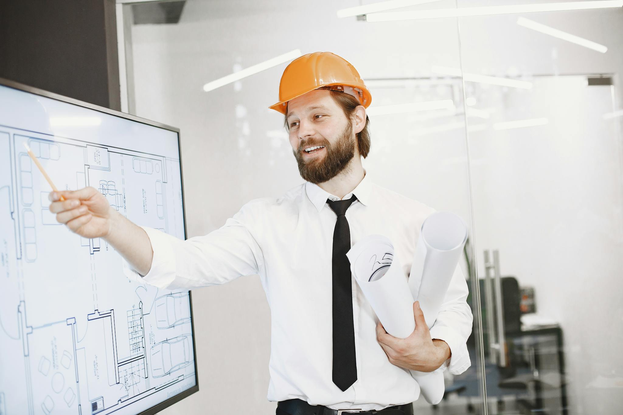 Engineer in helmet presenting architectural plans on a whiteboard in an office setting.